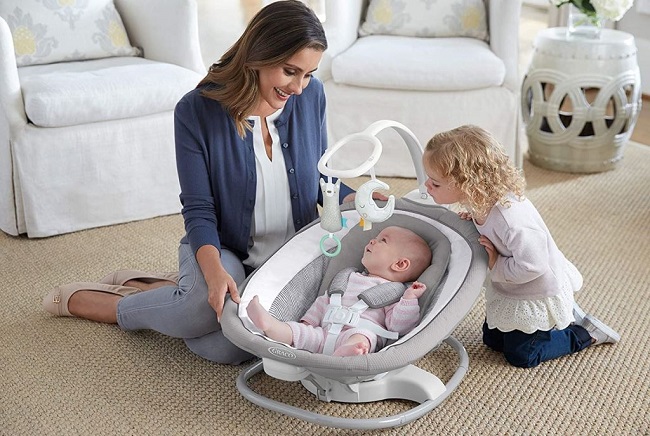 picture of a woman and a little girl sitting on a carpet in the living room, beside a baby in a bouncer swing