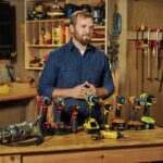 Man standing in a woodworking workshop with a collection of cordless power drills displayed on the workbench.