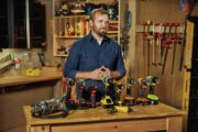 Man standing in a woodworking workshop with a collection of cordless power drills displayed on the workbench.
